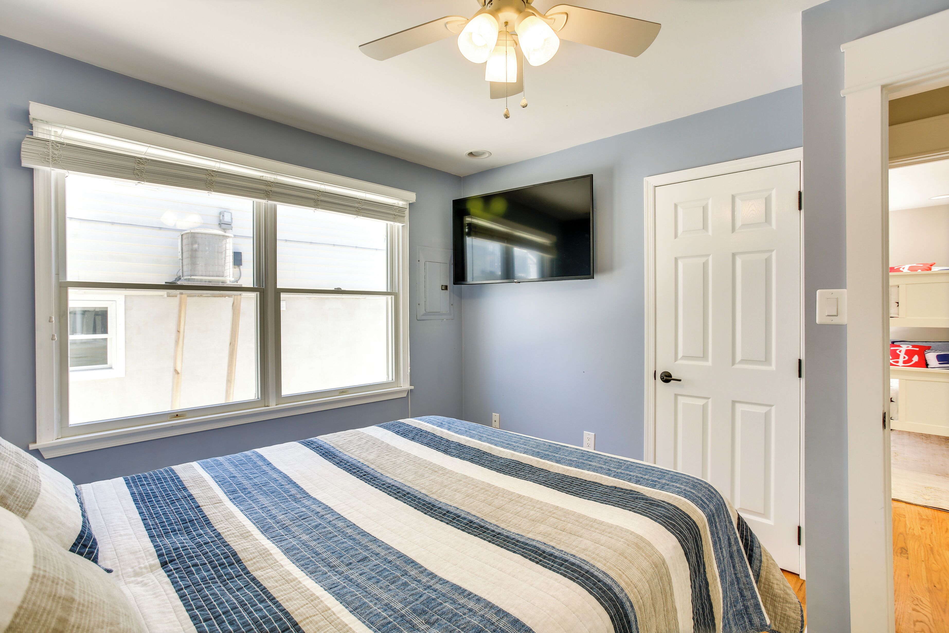 A bedroom with a large window, a ceiling fan, a flat-screen TV, and a door leading to another room.