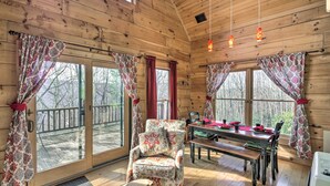 Interior de una cabaña de madera con comedor, un sillón mullido y ventanas grandes con cortinas.
