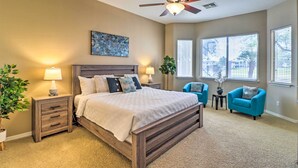 A bedroom with a large bed, two bedside tables with lamps, a ceiling fan, and a window with blinds. - Pool & Putting Green: Goodyear Home on Golf Course (Goodyear)
