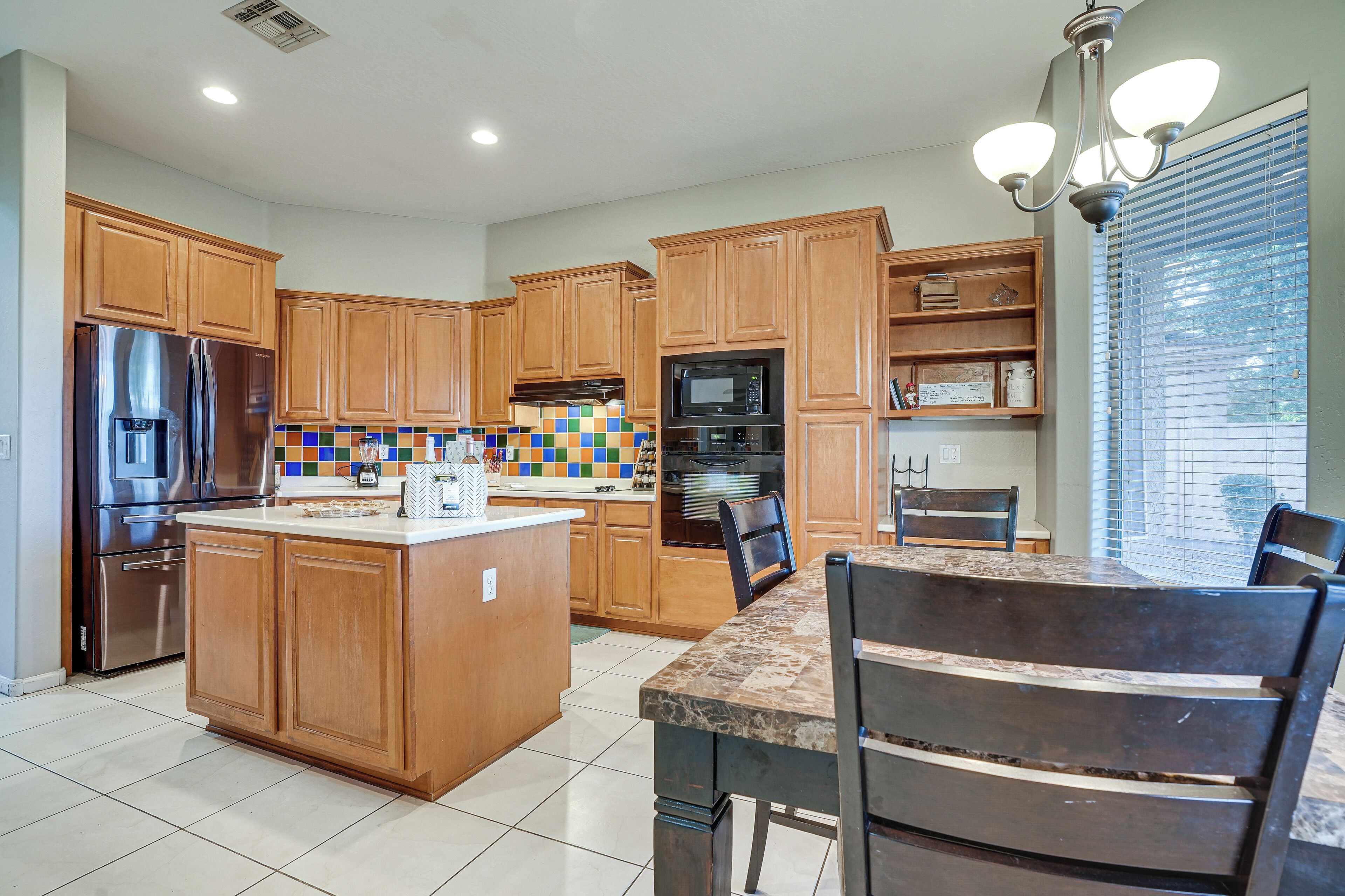 A kitchen with wooden cabinets, a central island, a dining table with chairs, and a window with blinds.