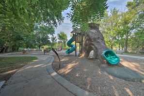 Ein Spielplatz mit Rutsche und Klettergerüsten, umgeben von Bäumen und einem gepflasterten Weg.