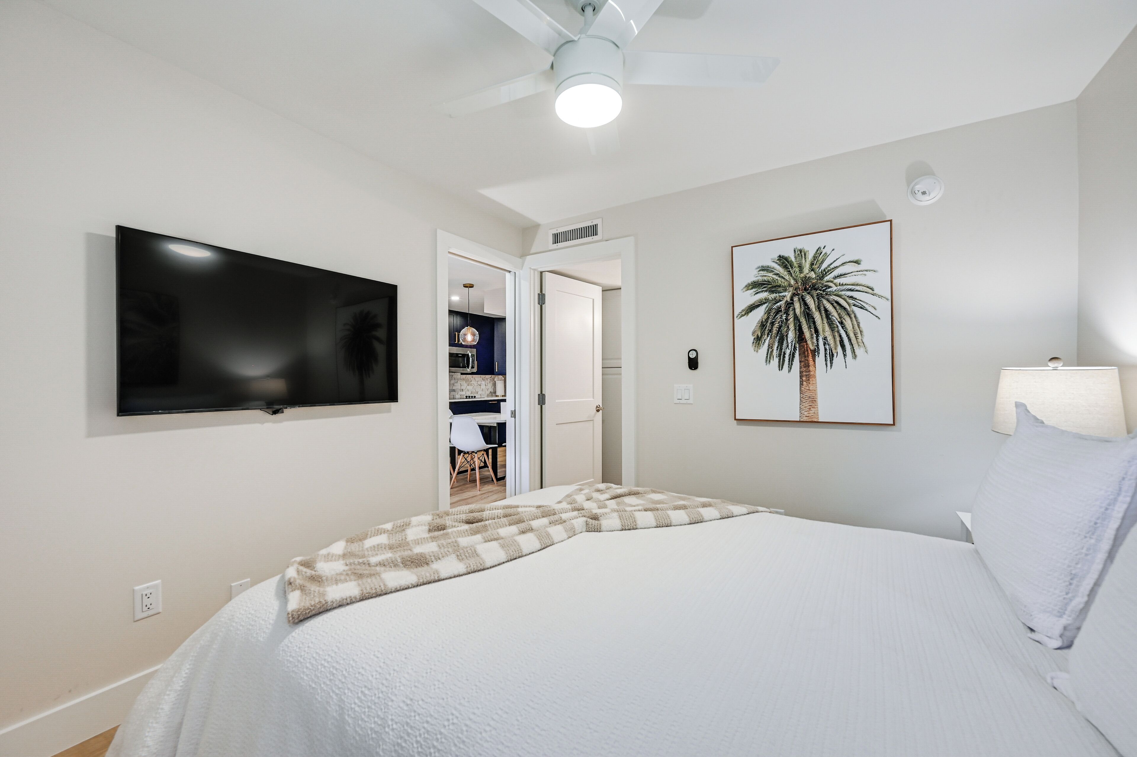 A bedroom with a bed, a TV mounted on the wall, a ceiling fan, and a framed picture of a palm tree.