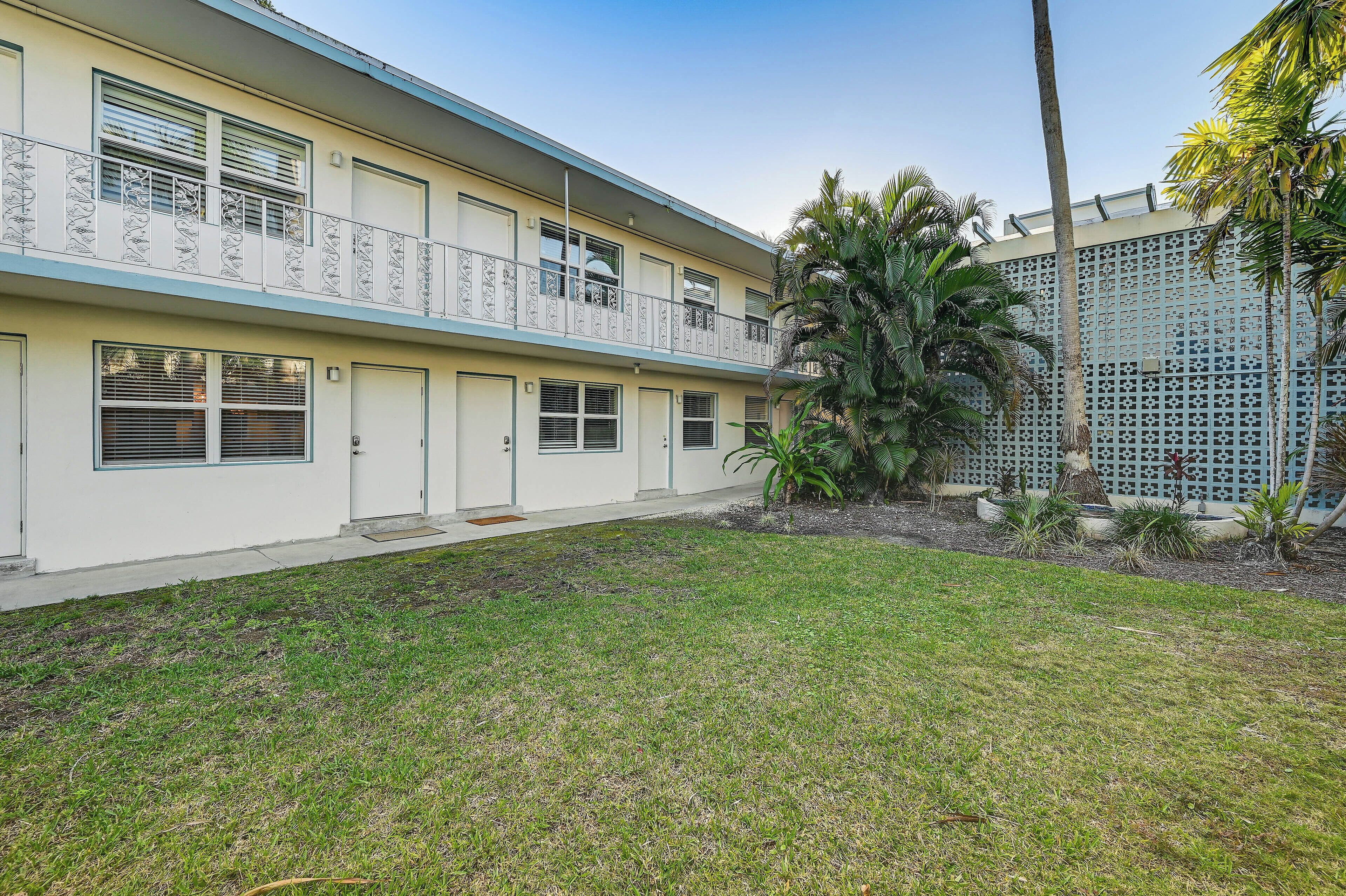 A two-story building with a balcony, a small garden area with palm trees, and a fence.