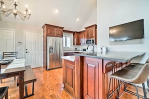 A modern kitchen with wooden cabinets, stainless steel appliances, a large island with bar stools, and a flat-screen TV mounted on the wall. - Relax Poolside! Family Getaway in Nashville Area (Pegram)