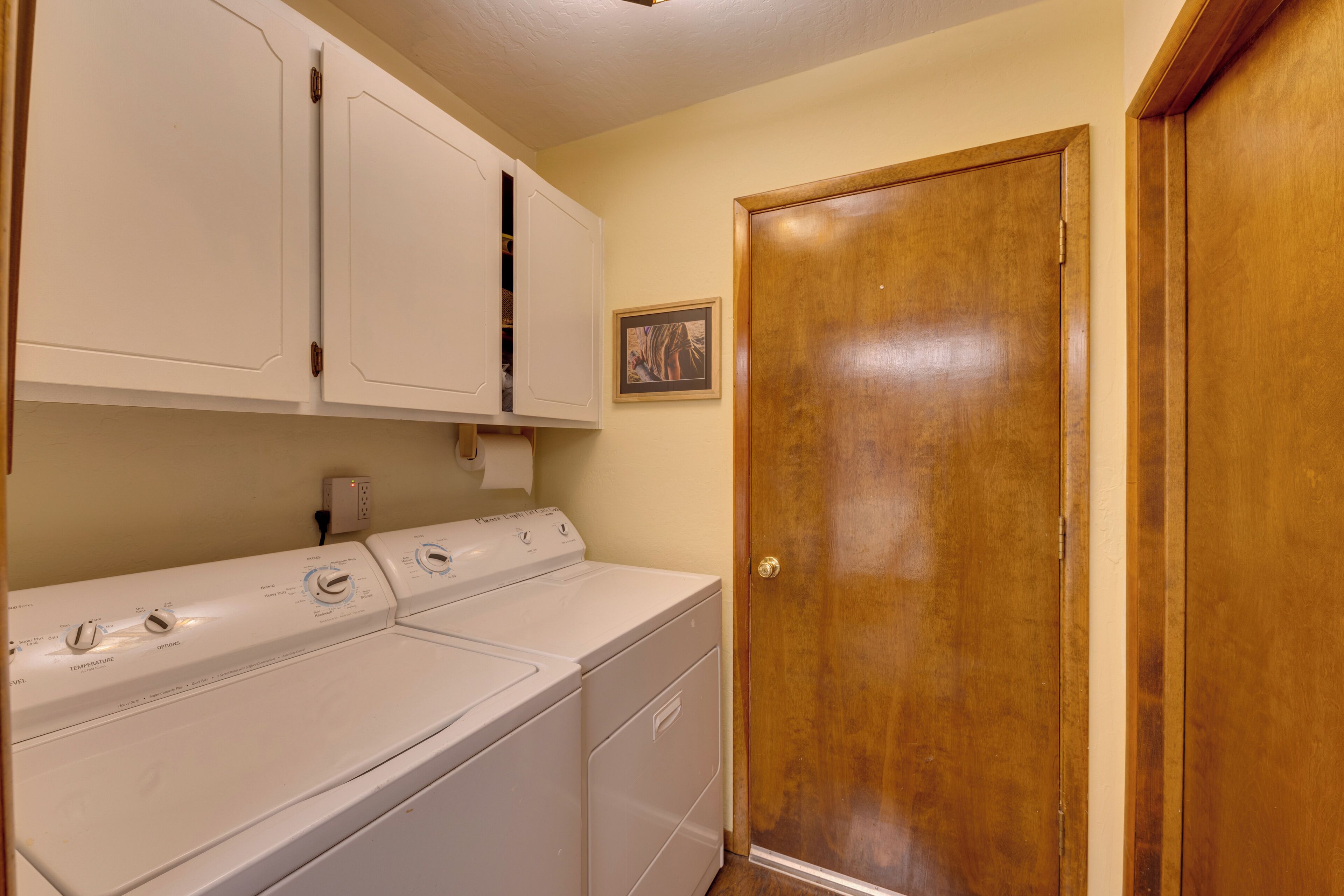A laundry area with a front-loading washer and dryer, wooden cabinets, a wooden door, and a small framed picture on the wall.
