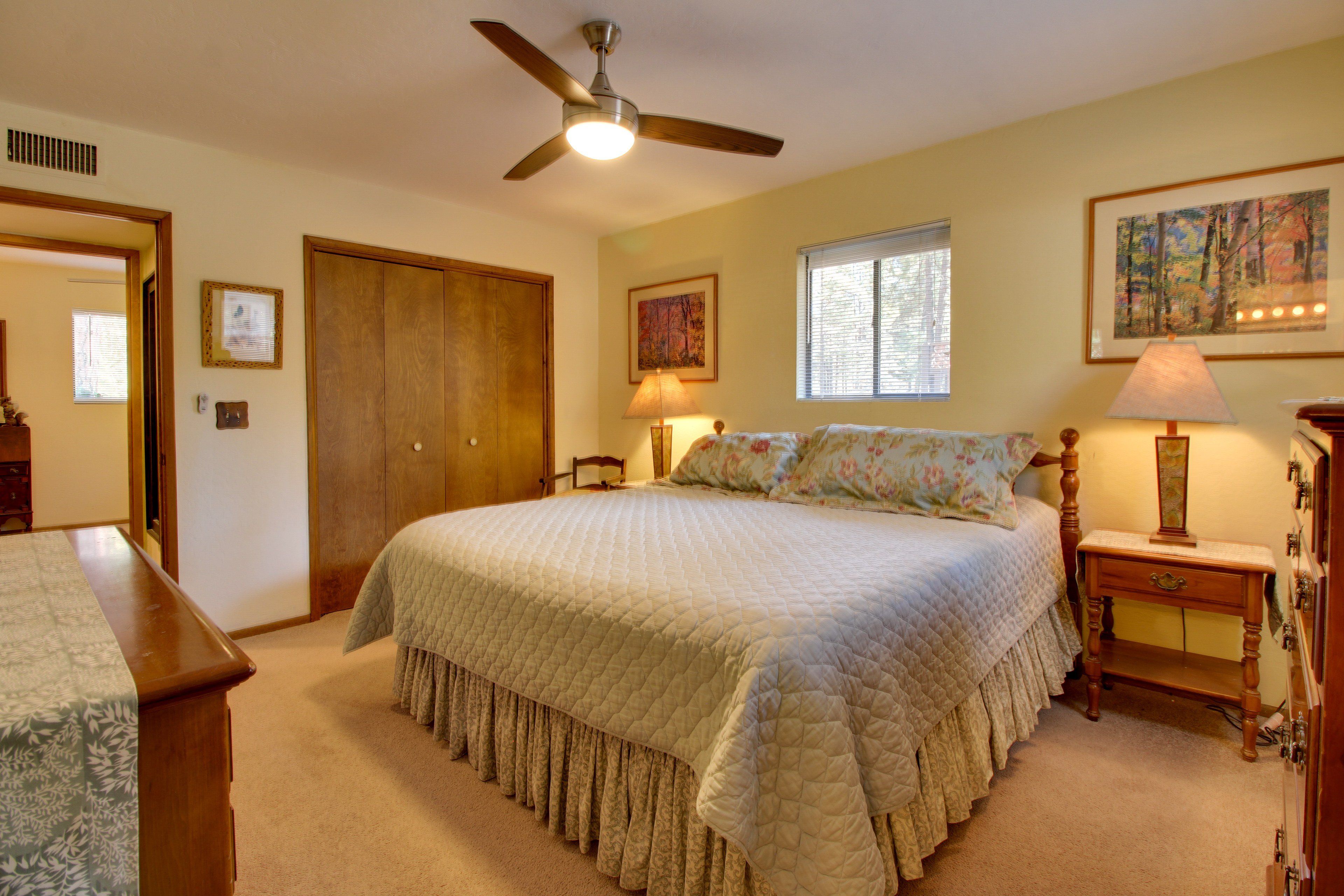 A bedroom with a large bed, a ceiling fan, a window with blinds, and a painting on the wall.
