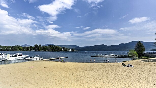 A beach with boats docked, a sandy shore, and mountains in the background. - Walk to Lake George! Bolton Landing Cottage (Bolton Landing)