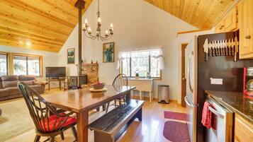 A spacious living room with a wooden ceiling, a dining area with a wooden table and benches, a kitchen with a refrigerator and knives hanging on it, and a fireplace.