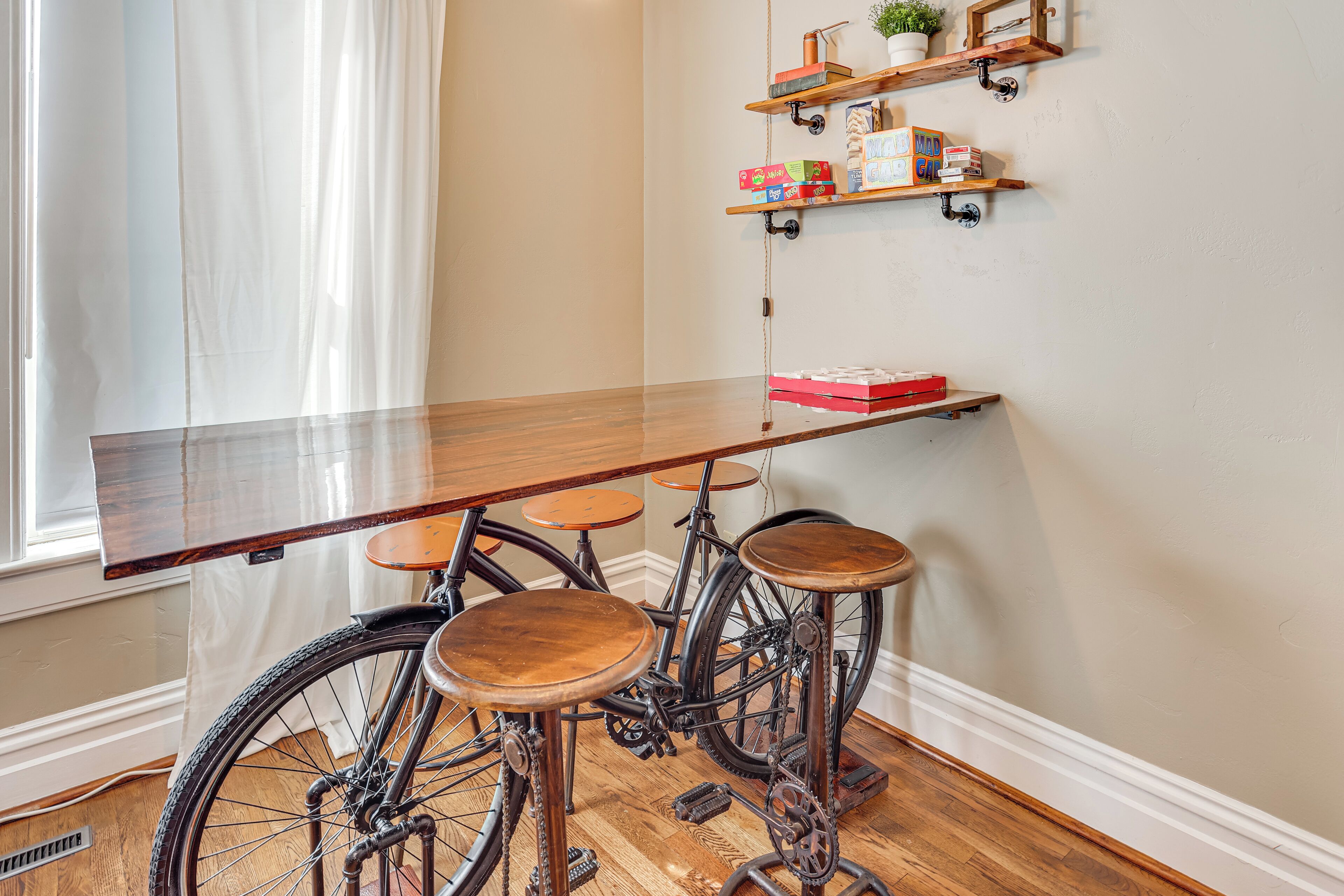 A wooden table with stools, a bicycle, and shelves with books and a plant.