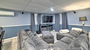 Un salon avec un canapé d’angle gris, un pouf gris, un téléviseur posé sur un meuble et une lampe.