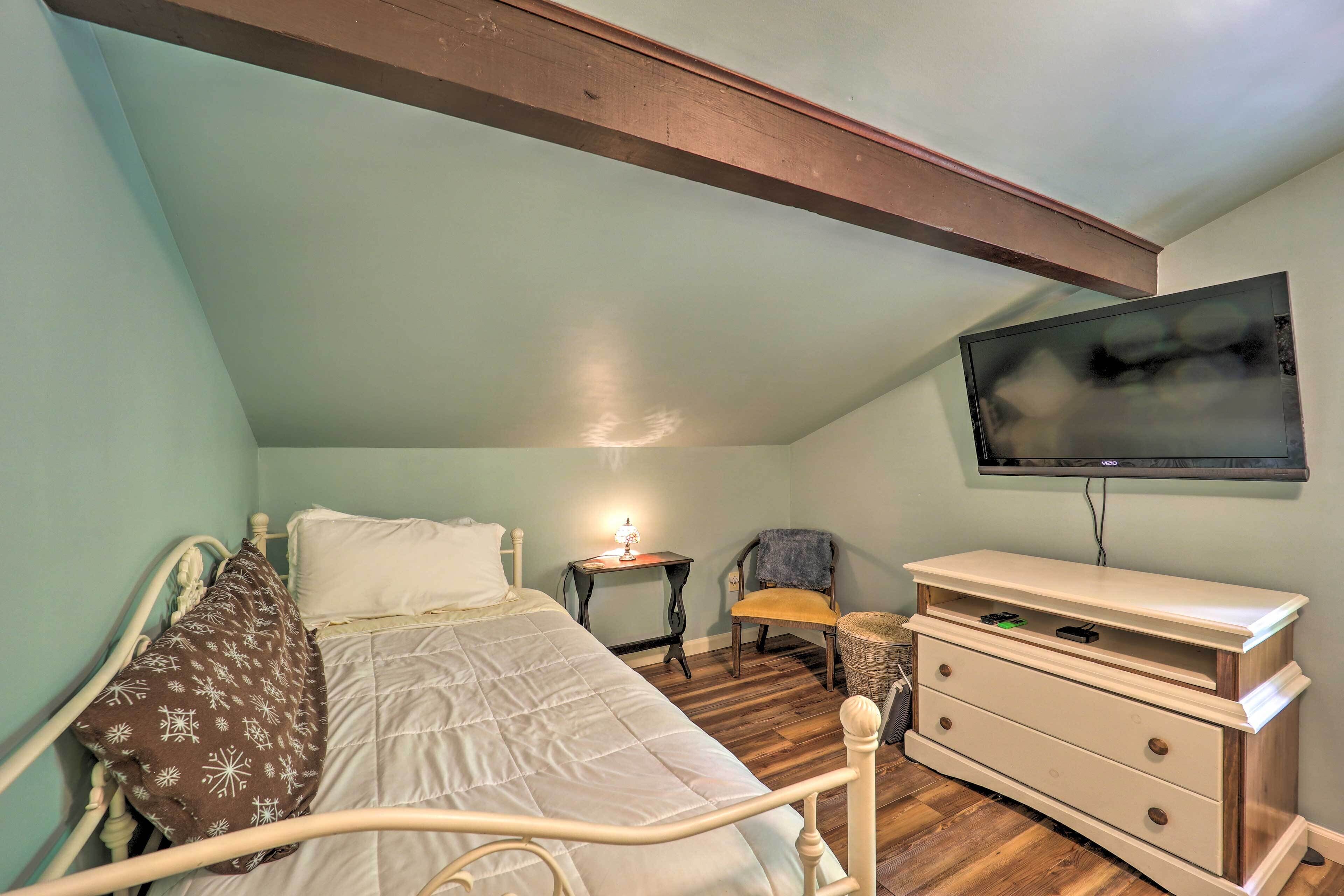A bedroom with a bed, a bedside table with a lamp, a chair, a TV mounted on the wall, and a dresser.