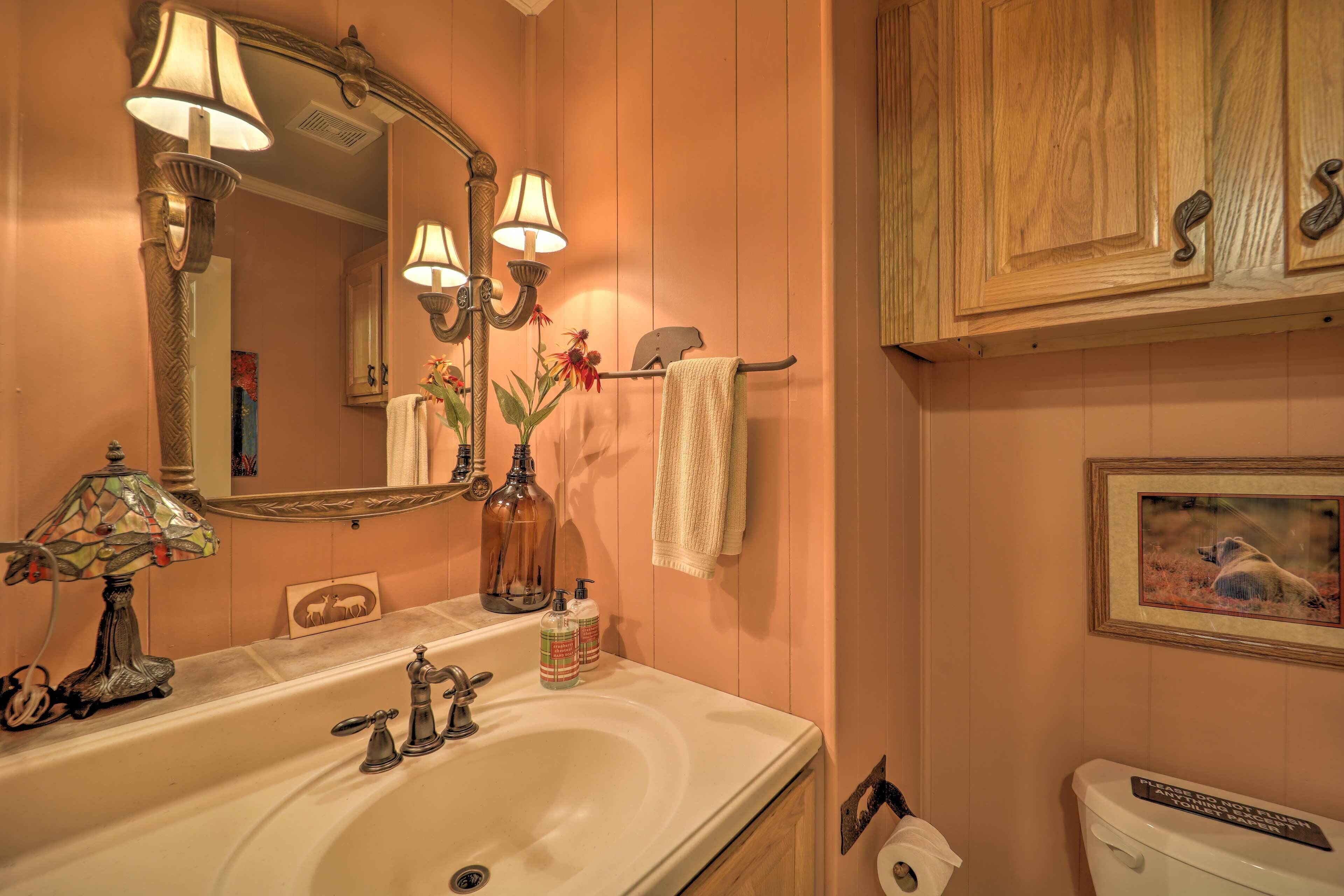 A bathroom with a large mirror, two wall-mounted lamps, a sink with soap dispensers, a towel rack, and a framed picture on the wall.