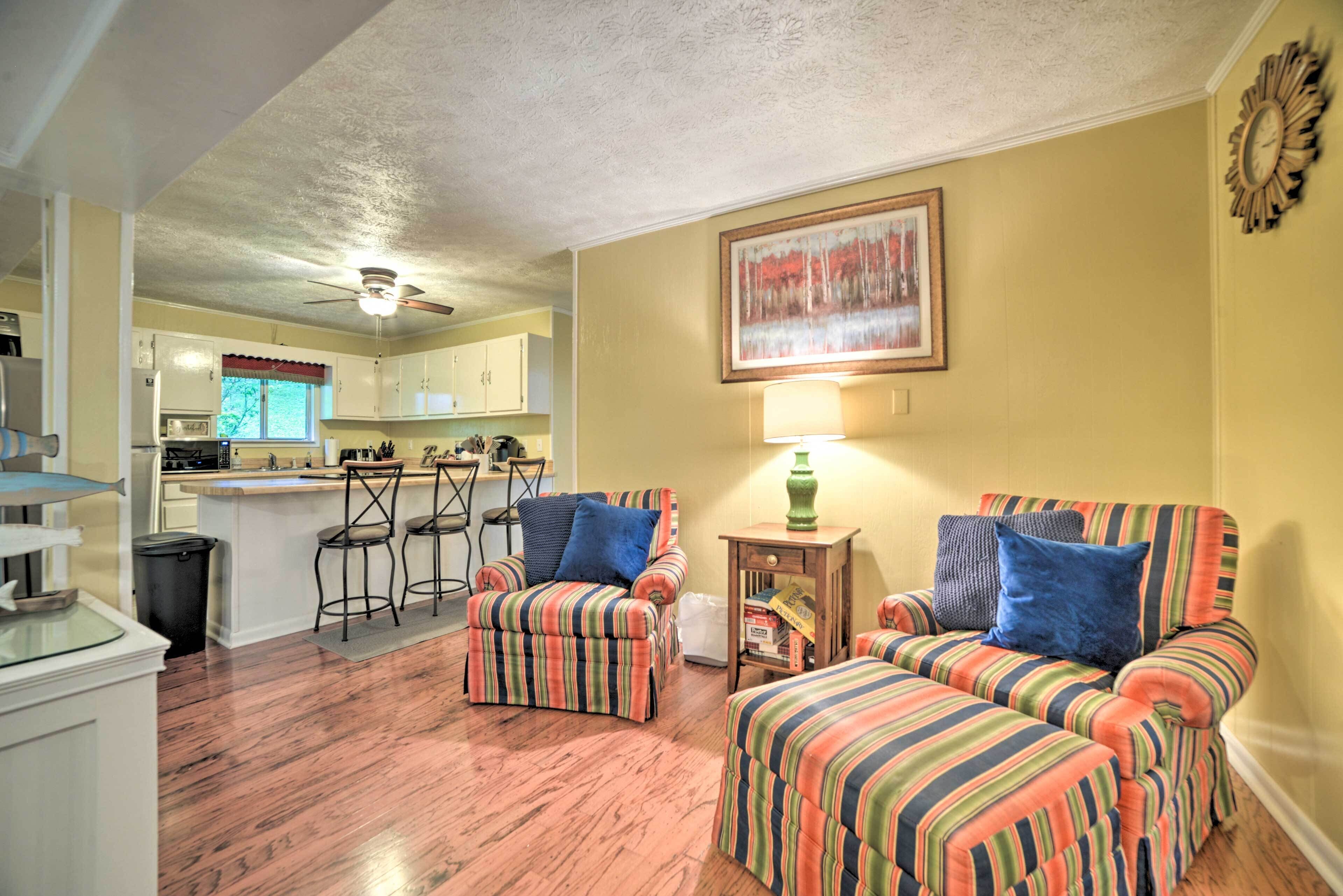 A living room with striped armchairs, a dining area with bar stools, and a kitchen in the background.
