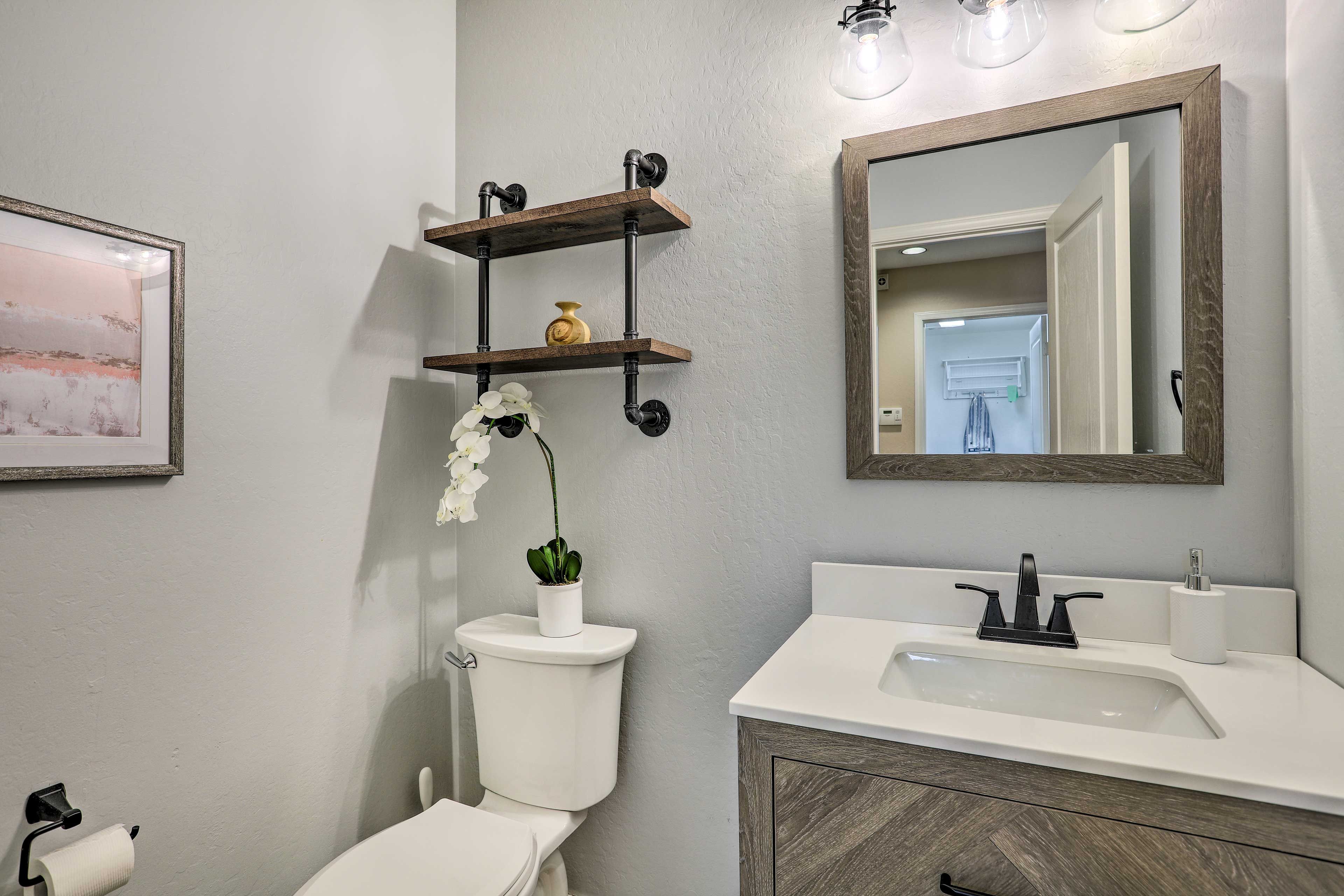 A bathroom with a toilet, sink, and a mirror. There are shelves above the toilet and a framed picture on the wall.
