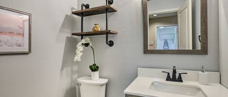 Un baño con inodoro, lavabo y espejo. Hay repisas sobre el inodoro y un cuadro en la pared.