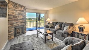 A living room with a stone fireplace, a gray sofa set, two side tables with lamps, and a view through a glass door to an outdoor area. - Scottsdale Condo w/ Pool Access: Hike, Swim & Shop (Scottsdale)