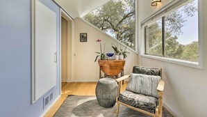 A modern interior with a wooden chair, a round ottoman, a wooden side table with plants, and a large window with a view of trees. - Luxe Santa Rosa Home ~ 3 Mi to Downtown! (Santa Rosa)