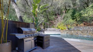 An outdoor kitchen area with a grill, oven, and storage drawers, surrounded by a wooden deck and a stone wall. - Luxe Santa Rosa Home ~ 3 Mi to Downtown! (Santa Rosa)