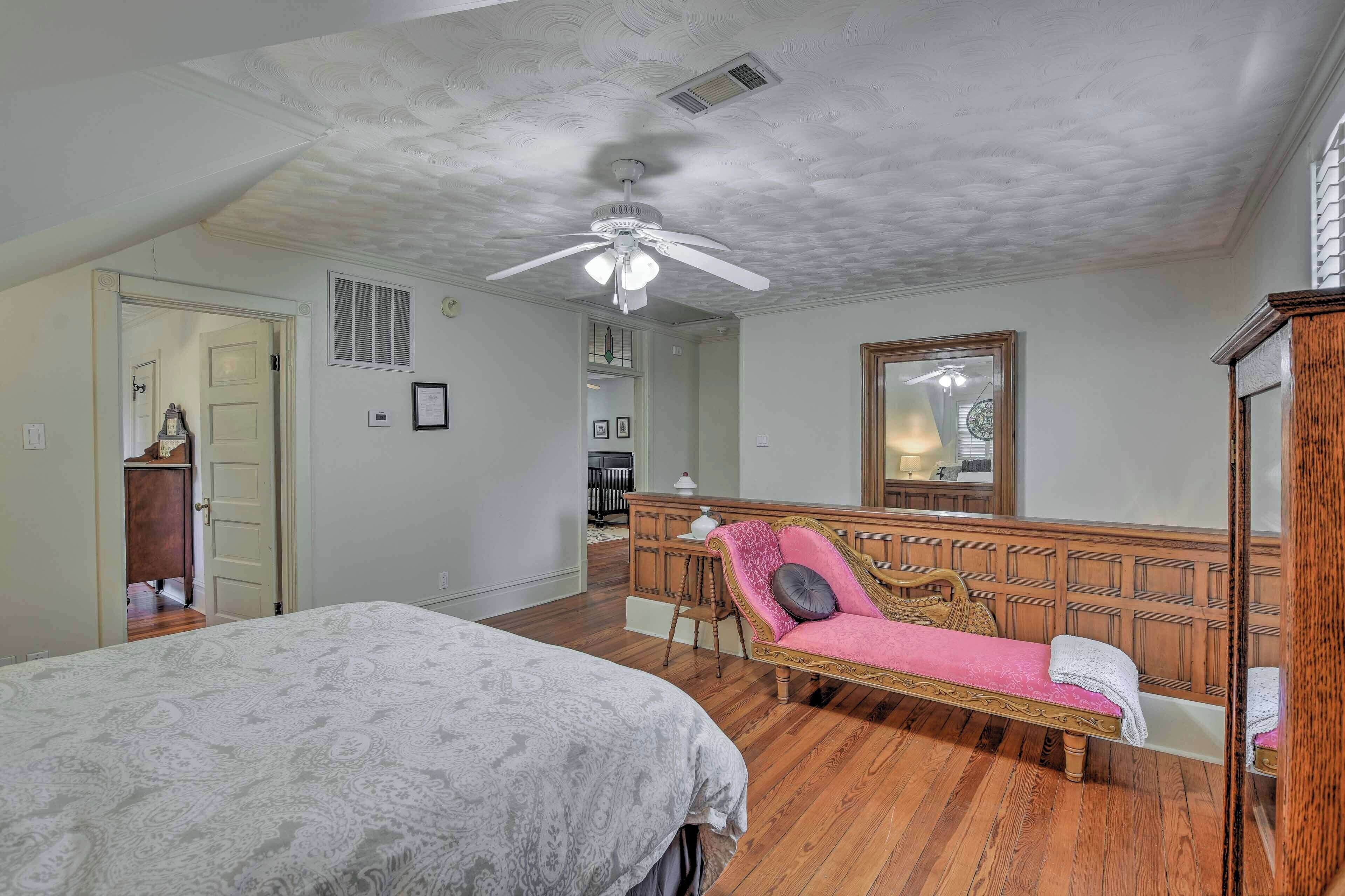 A bedroom with a bed, a pink chaise lounge, a wooden dresser, and a ceiling fan.