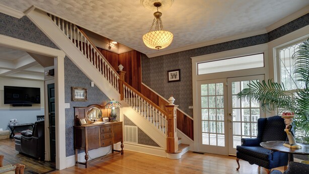 A spacious living room with hardwood floors, a wooden staircase, a chandelier, and a large window with a view. - Historic Hot Springs Home < 1 Mi to Oaklawn! (Hot Springs)