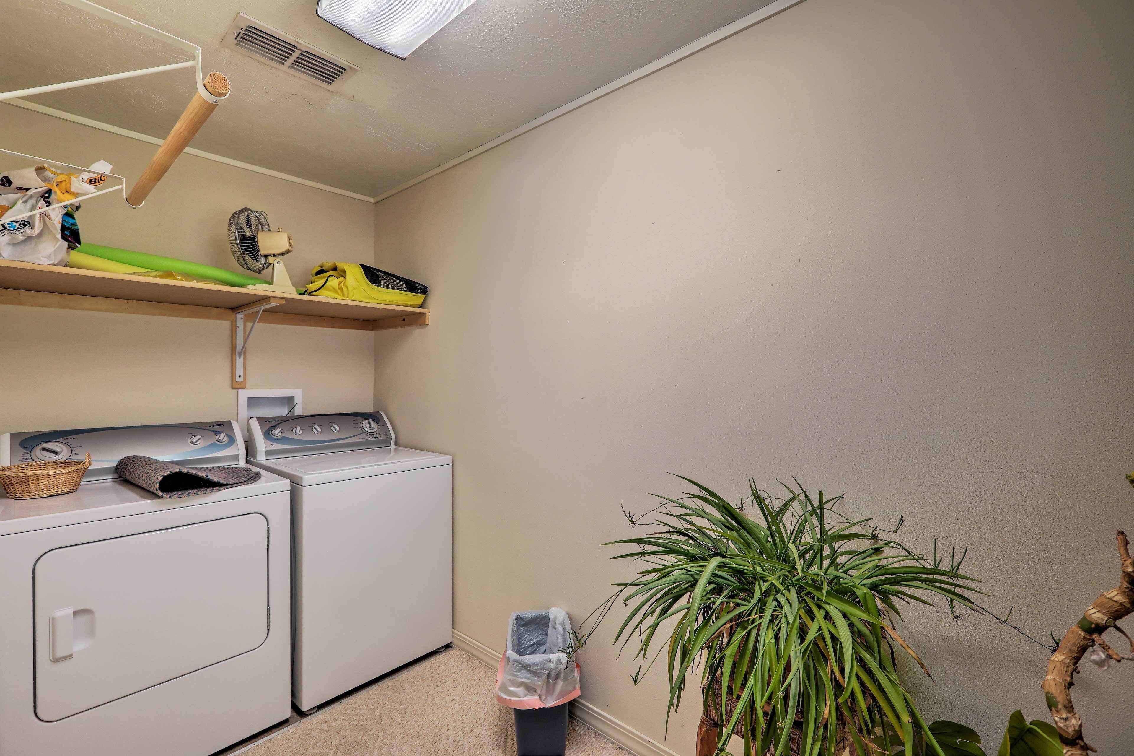 A laundry room with two white washing machines, a shelf with various items, a fan, and a potted plant.