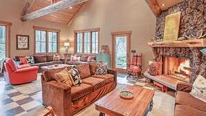 A spacious living room with a high ceiling, a fireplace, and a large window overlooking a forested area.