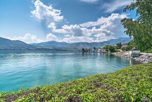 A serene lakeside view with mountains in the background, a dock extending into the water, and a small building on the shore. - Lake Chelan Condo, Walk to Brewery & Wineries (Manson)