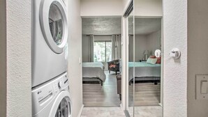 Une buanderie équipée d’un lave-linge et d’un sèche-linge à chargement frontal, un couloir menant à une chambre avec un lit et une table de chevet, et une fenêtre avec des rideaux.