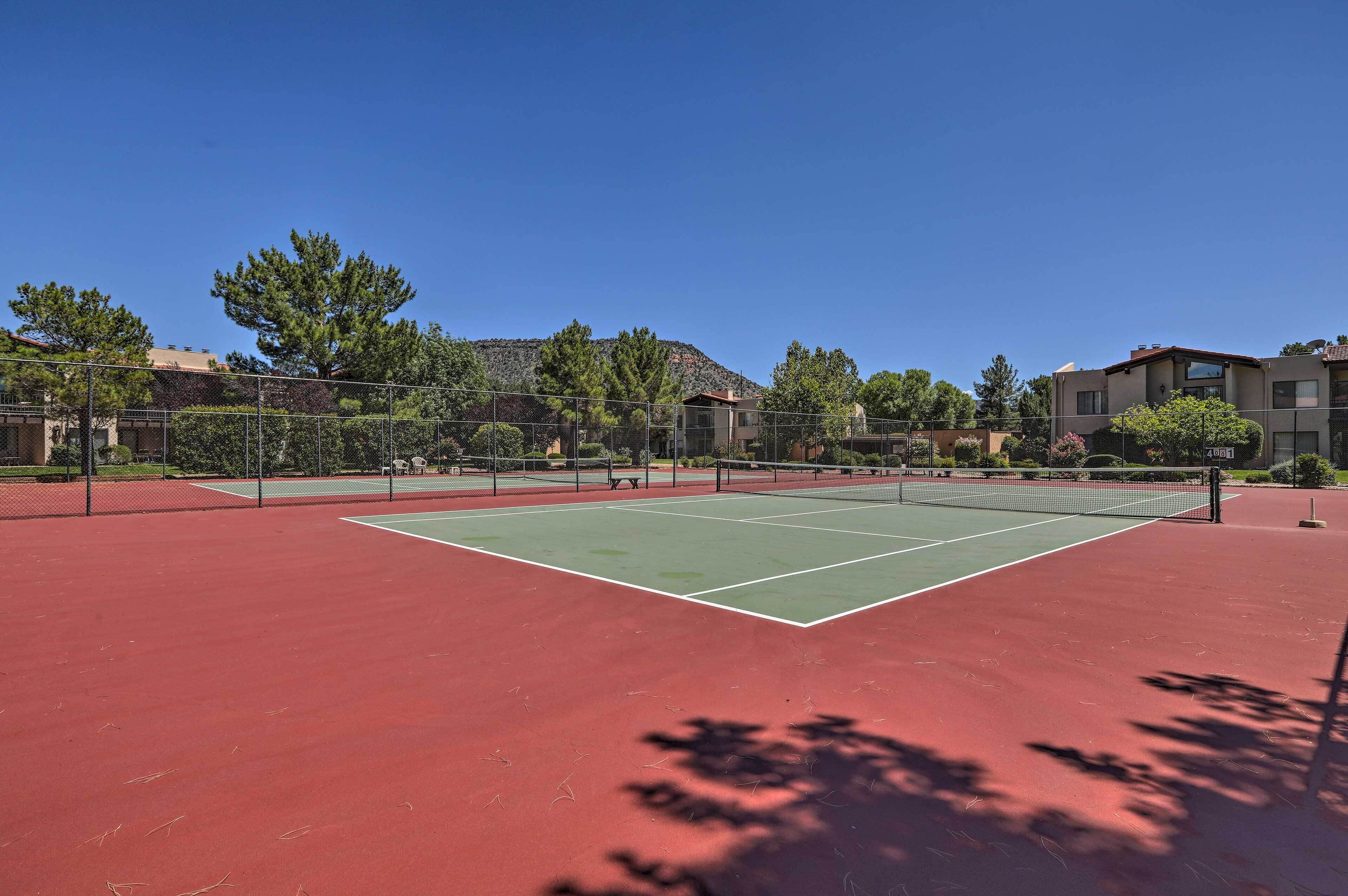 A tennis court with a red and green surface, surrounded by trees and buildings.