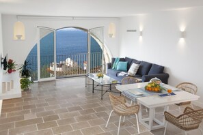 A spacious balcony with a view of the sea, furnished with a sofa, chairs, and a table with fruits and a drink.