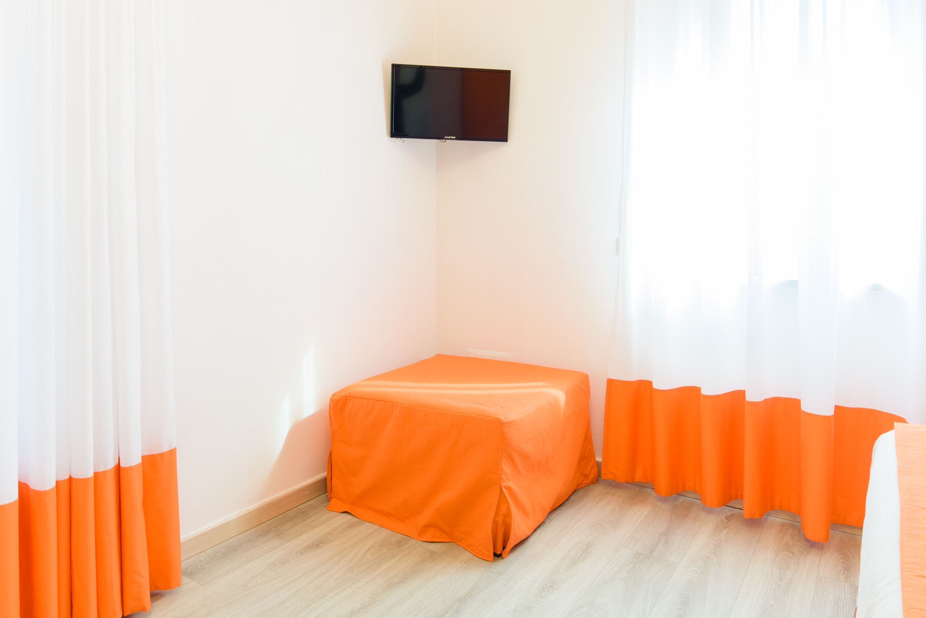 Ein Zimmer mit einem kleinen orangen Tisch, einem flachbildfernseher an der Wand und transparenten Vorhängen mit orangen Akzenten.