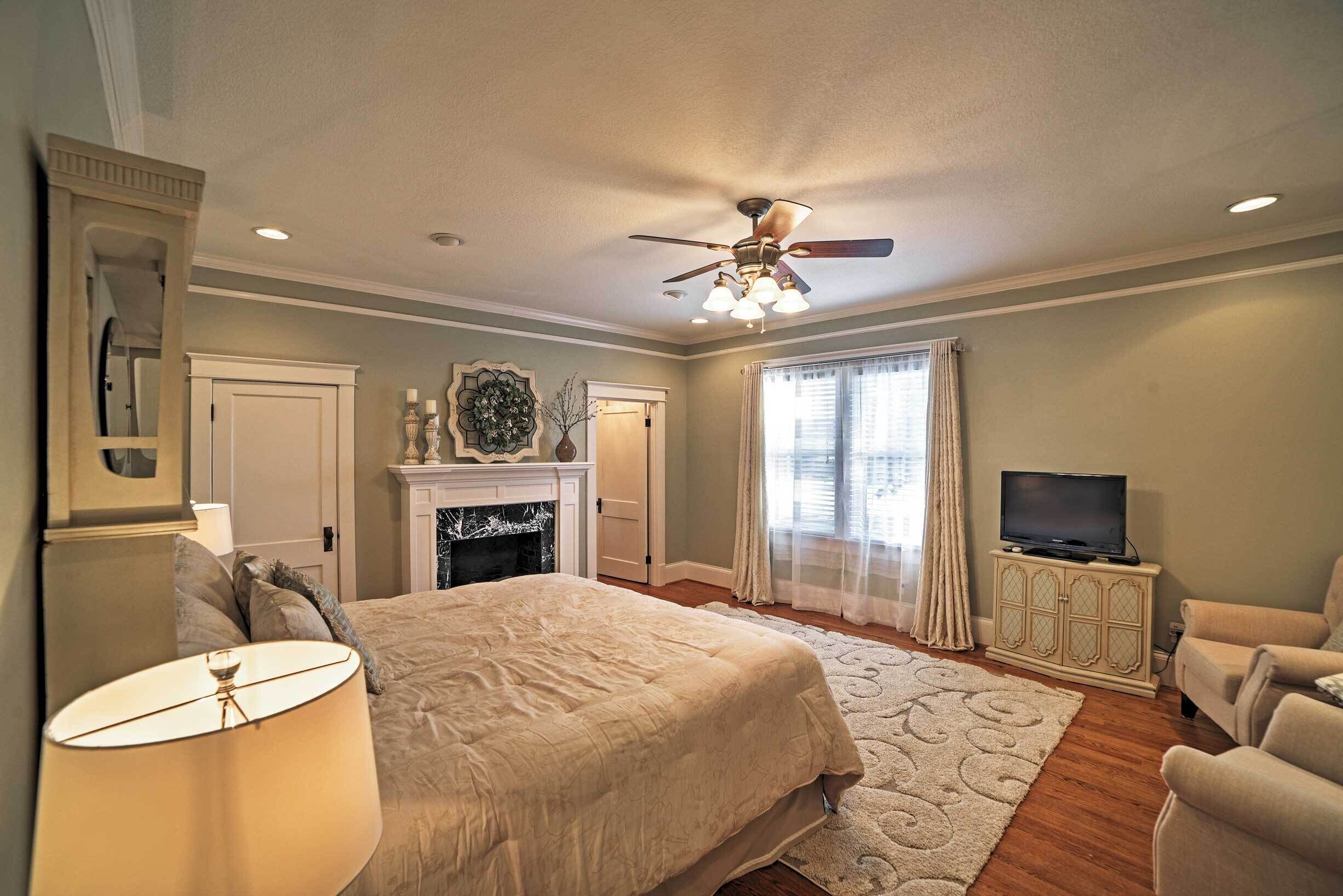 A bedroom with a bed, a ceiling fan, a fireplace, a television on a cabinet, and a window with curtains.