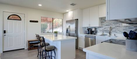 A modern kitchen with white cabinetry, a large island with bar stools, stainless steel appliances, and a view of the outdoors.