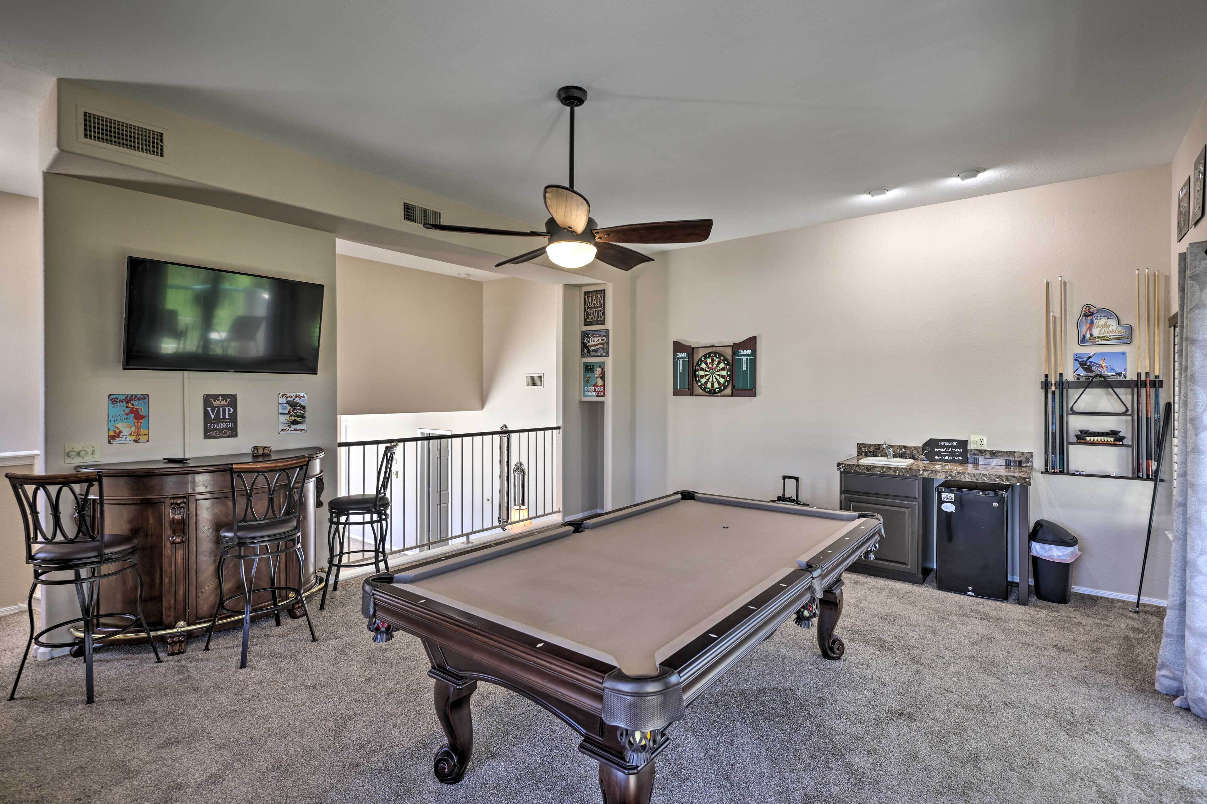 A pool table in a room with a ceiling fan, a bar area with stools, a dartboard, and a television mounted on the wall.