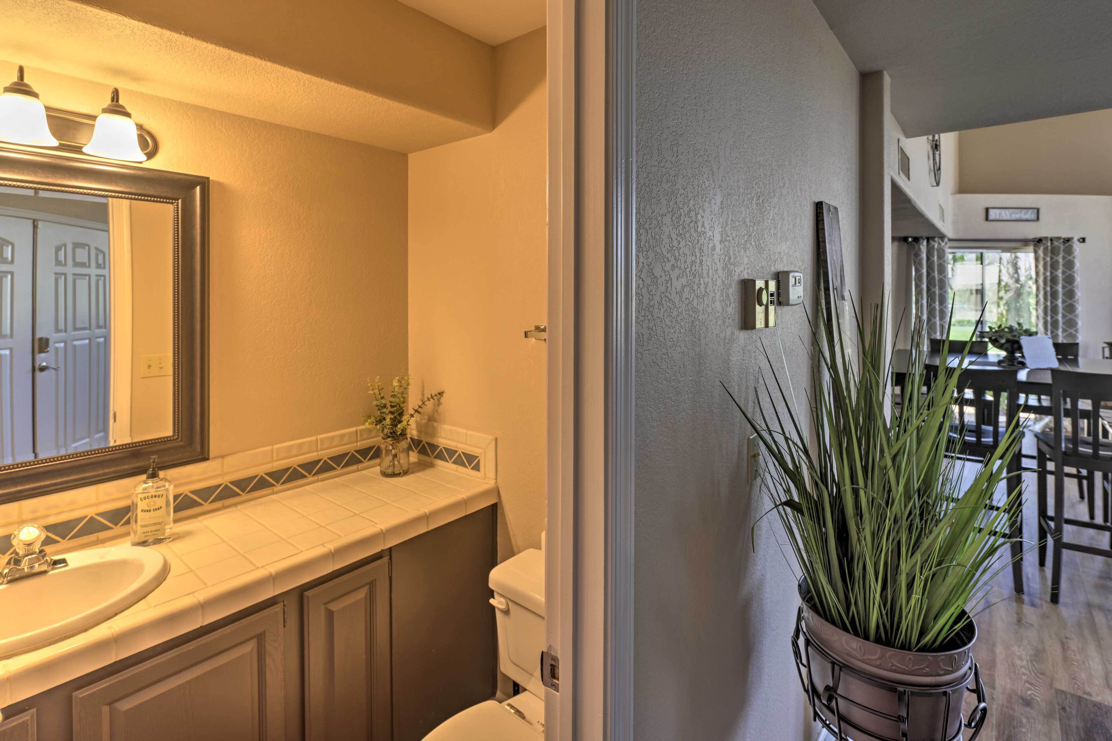 Een badkamer met een wastafel, spiegel en toilet. Er staat een plant in een pot aan de muur en een deur die leidt naar een andere kamer.