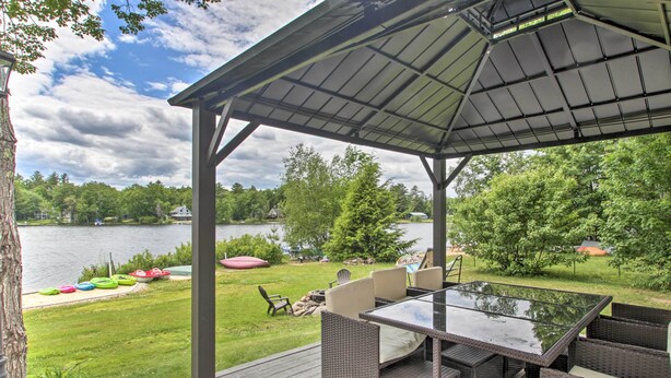 A covered outdoor patio with a glass-top table and wicker furniture, overlooking a lake and greenery. - New Hampshire Home w/ Private Beach, Dock & Rafts! (Center Barnstead)