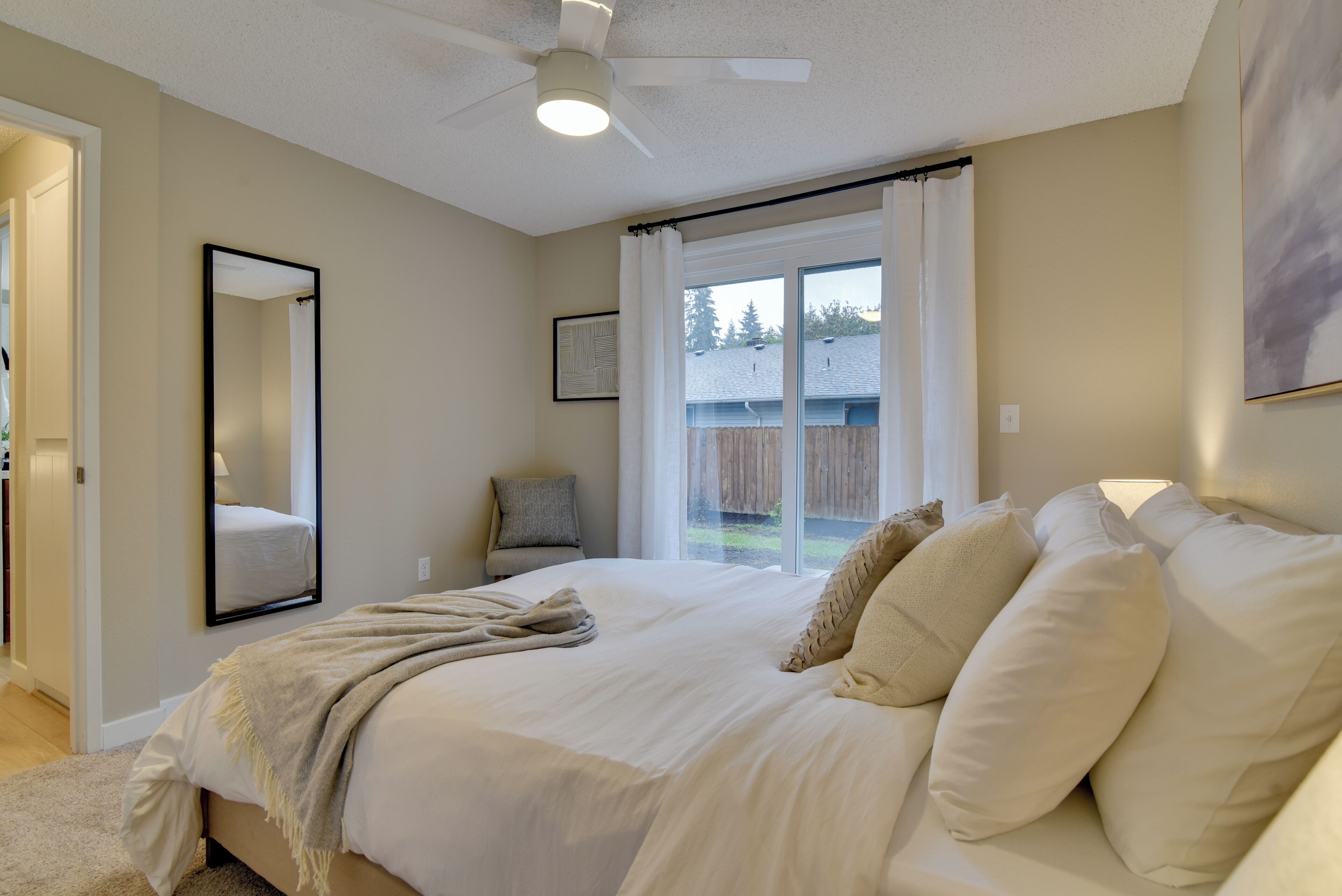 A bedroom with a large bed, a ceiling fan, a window with curtains, a mirror, and a door leading to another room.