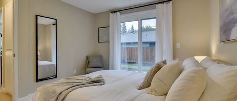 A bedroom with a large bed, a ceiling fan, a window with curtains, a mirror, and a door leading to another room.