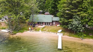An aerial view of a cabin with a green roof, surrounded by trees and a sandy beach. - 13 Mi to Dtwn: Lakefront Cottage w/ Dock (Traverse City)