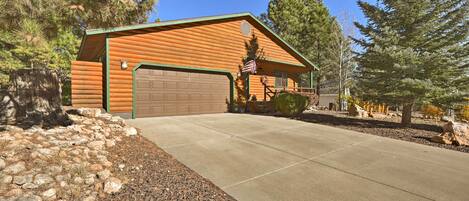 A log cabin with a garage, surrounded by trees and a gravel driveway.