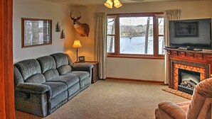 A cozy living room with a fireplace, a mounted deer head, a television, and a large window overlooking a lake. - Cozy Balsam Lake Home: Deck, Private Dock + Kayaks (Balsam Lake)