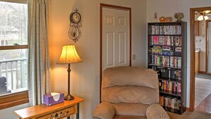 A living room with a beige armchair, a wooden side table with a lamp, a bookshelf filled with books, and a door with a clock on the wall. - Cozy Balsam Lake Home: Deck, Private Dock + Kayaks (Balsam Lake)