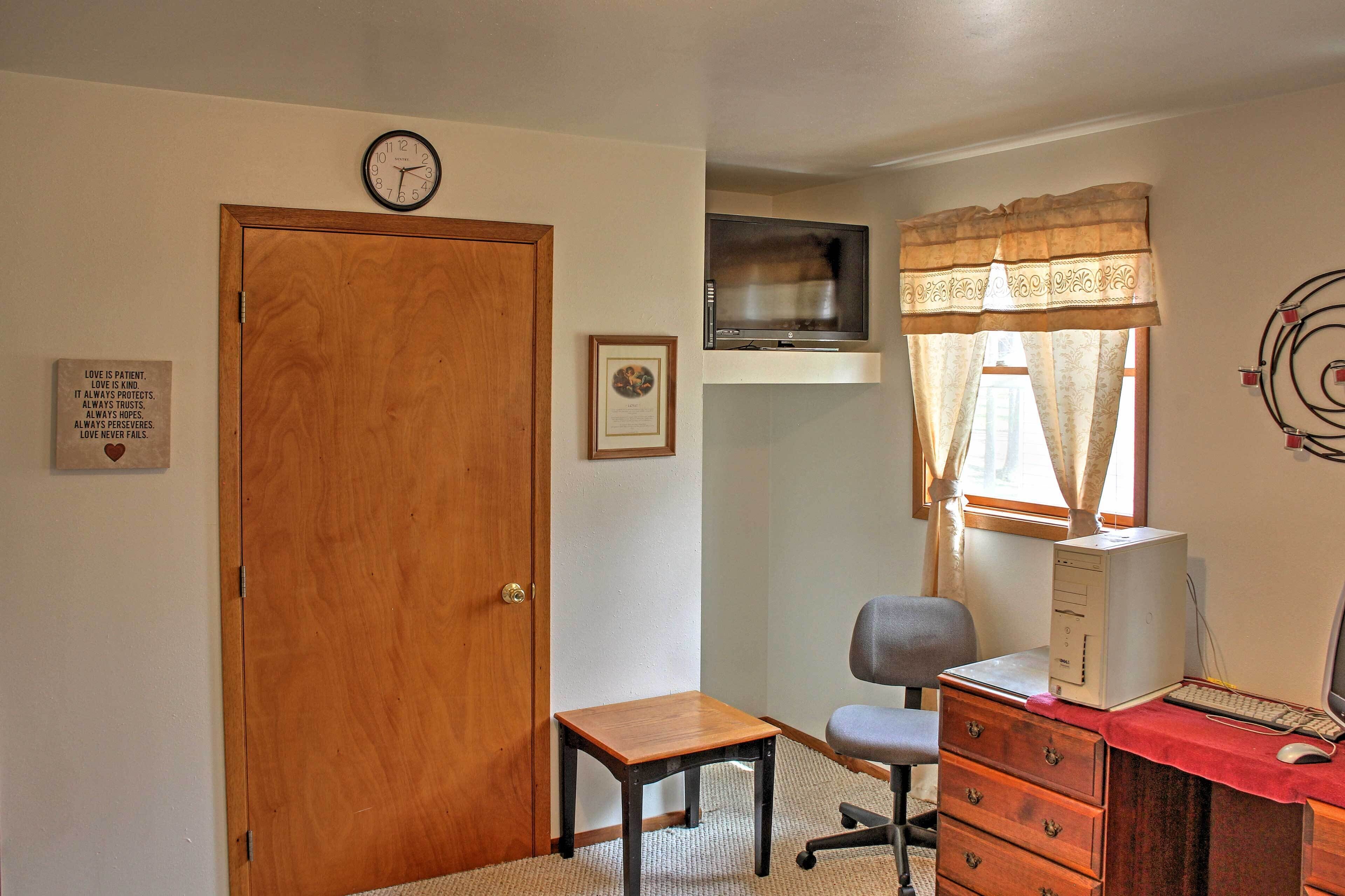 A room with a wooden door, a small table, a chair, a dresser, a desk with a computer, a TV, and a clock on the wall.