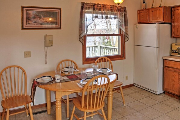 Comedor con mesa de madera preparada para cuatro personas, rodeada de sillas de madera. Se ve una ventana con vista a una terraza y árboles, y un refrigerador blanco al fondo.
