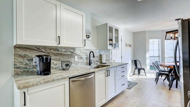 A modern kitchen with white cabinetry, a marble countertop, and a dishwasher. There is a dining area with a table and chairs visible in the background.