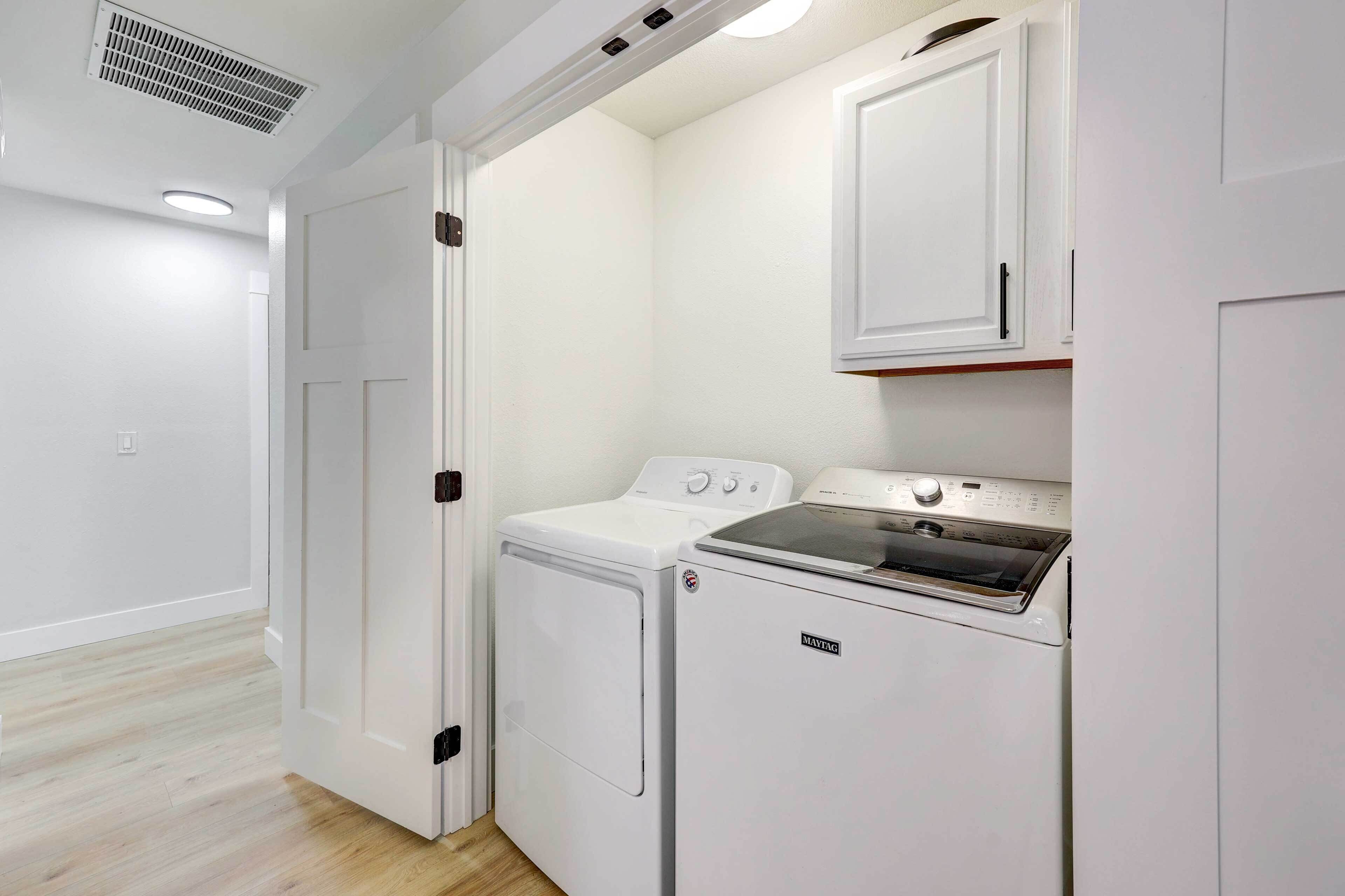 Una sala de lavandería con lavadora y secadora de carga frontal, un armario sobre la secadora y una puerta que da a otra habitación.
