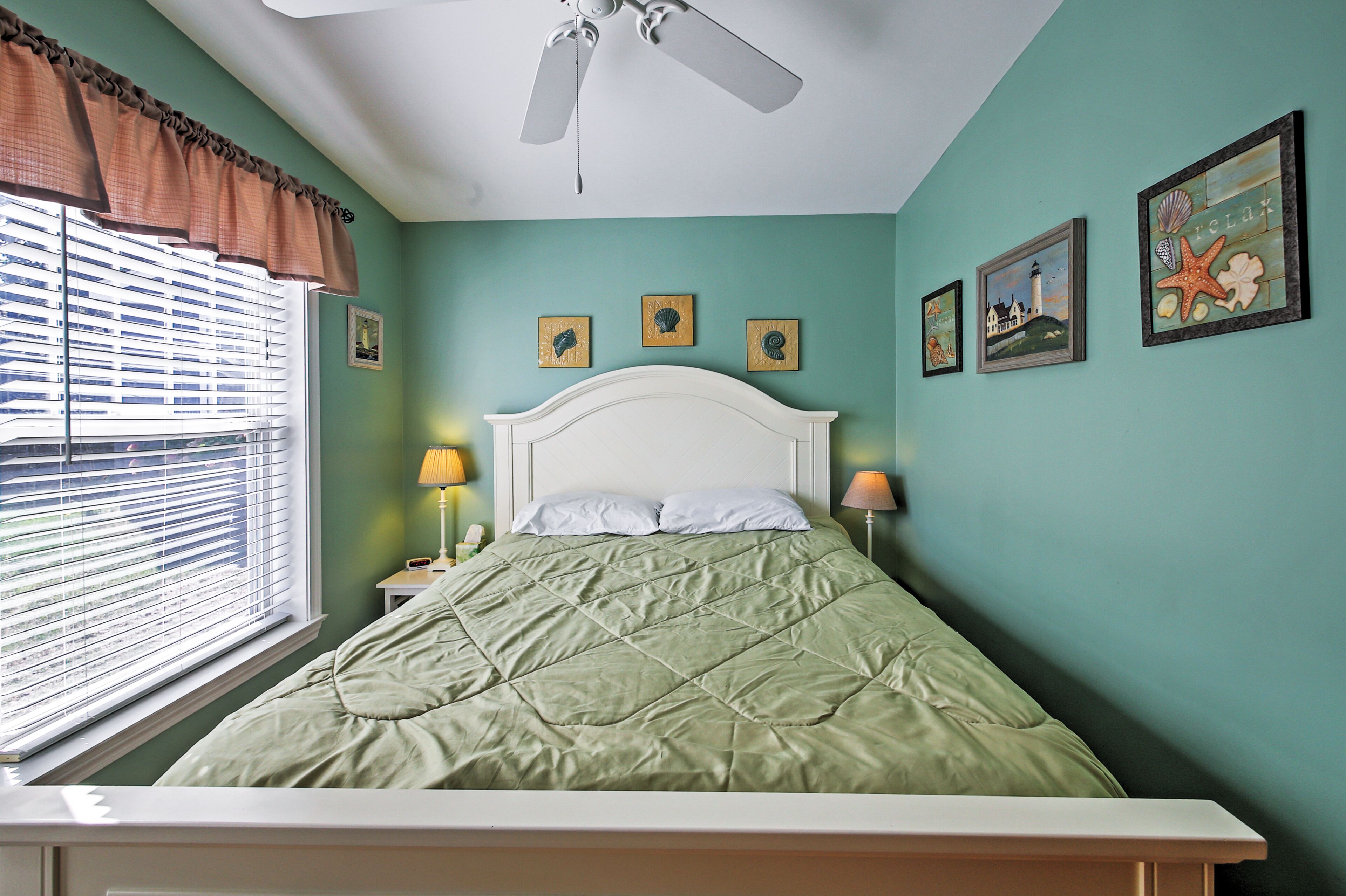 Une chambre à coucher comprenant un lit, un ventilateur de plafond, une fenêtre avec des stores et des œuvres d’art murales.