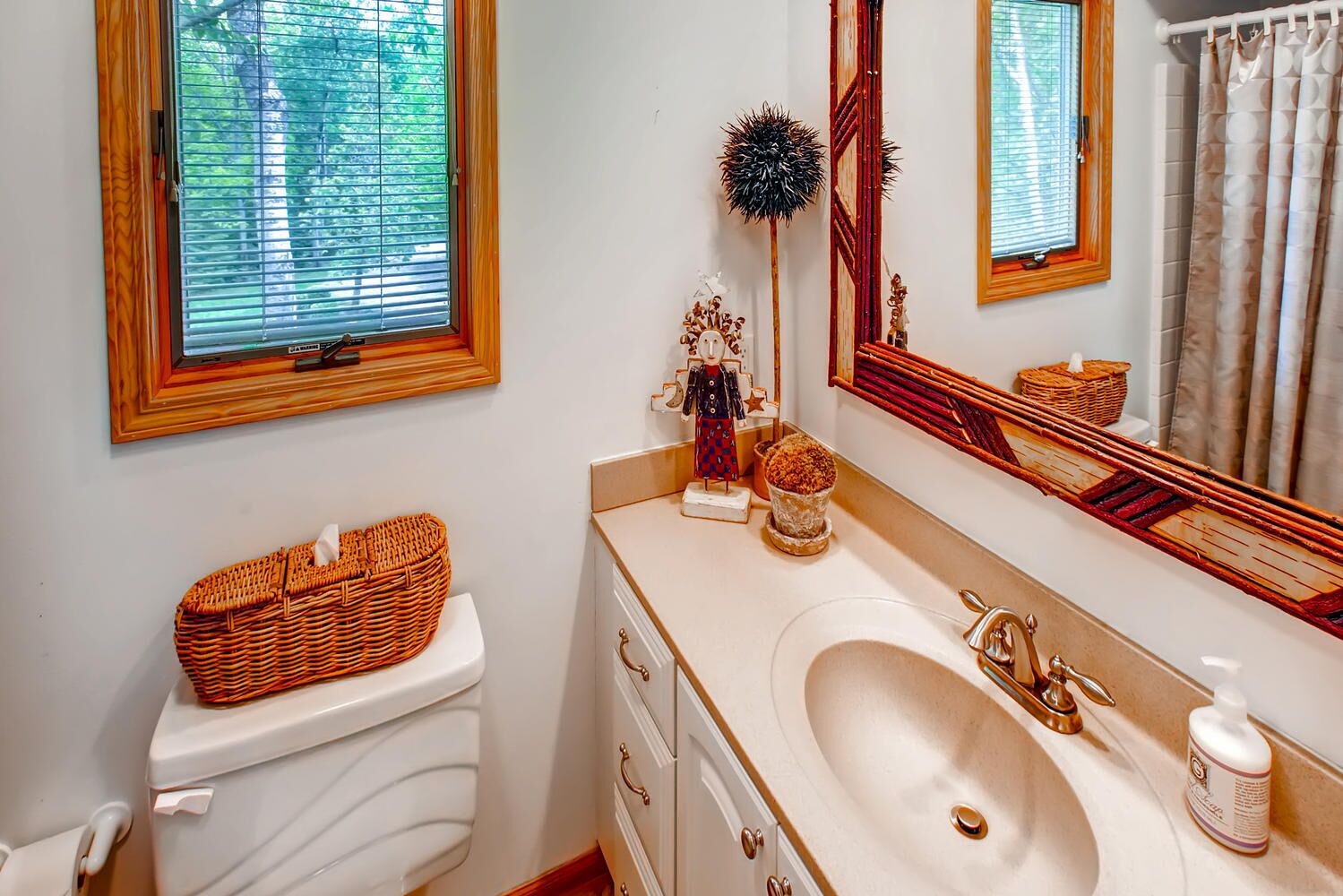 Une salle de bain avec des toilettes blanches, un lavabo beige avec un robinet doré, un grand miroir et un panier en osier posé sur les toilettes.