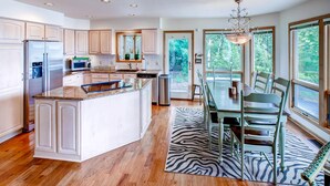 A modern kitchen with white cabinetry, a large island, and a dining area with a zebra-patterned rug. - Lavish Charlevoix Home on Lake Michigan, Fire Pit (Charlevoix)
