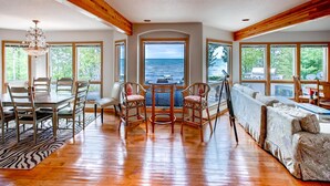 A spacious room with a wooden floor, large windows offering ocean views, a dining area with chairs and a table, and a sofa. - Lavish Charlevoix Home on Lake Michigan, Fire Pit (Charlevoix)